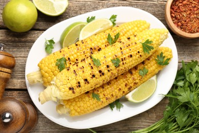 Photo of Tasty grilled corncobs with parsley and lime slices on wooden table, flat lay