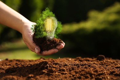 Image of Electricity and nature. Woman holding soil with green plant and light bulb outdoors, closeup