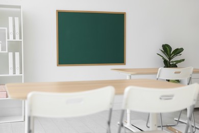 Photo of Blank green chalkboard, desks, chairs and plant in classroom