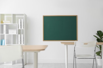 Photo of Blank green chalkboard, desks, chairs, shelving unit and plant in classroom