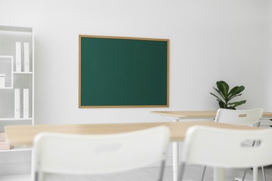 Photo of Blank green chalkboard, desks, chairs, shelving unit and plant in classroom