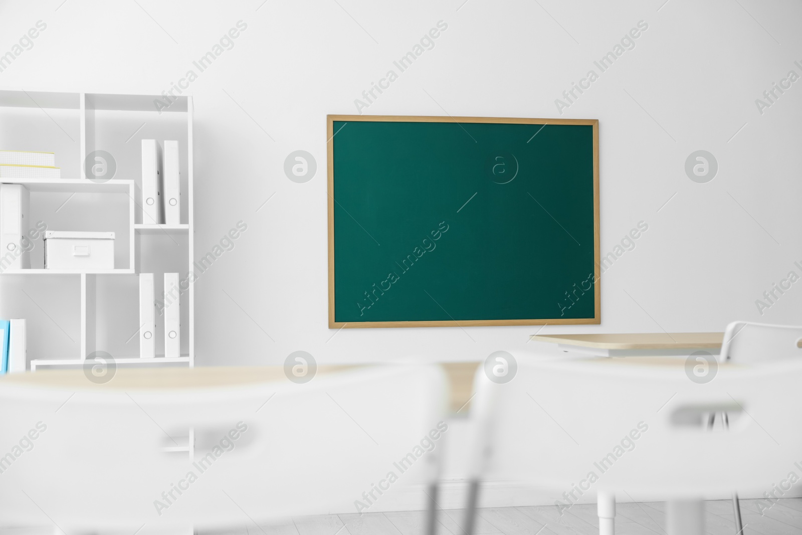 Photo of Blank green chalkboard, desks, chairs and shelving unit in classroom