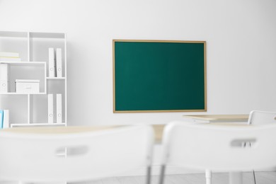 Photo of Blank green chalkboard, desks, chairs and shelving unit in classroom