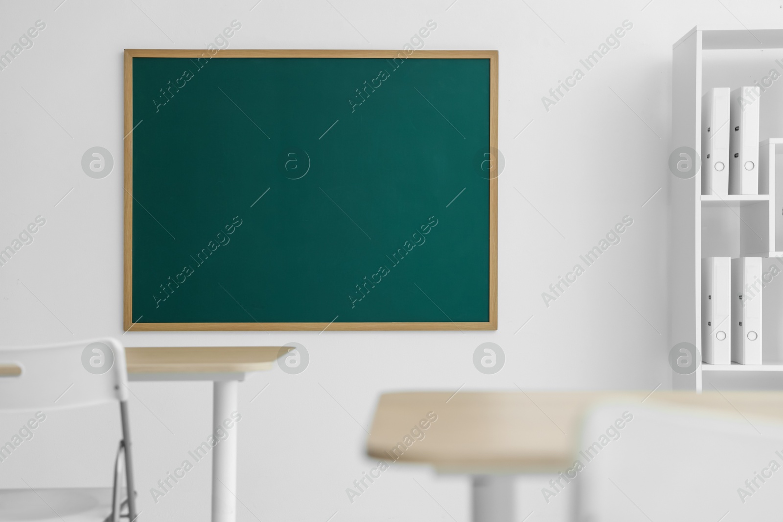 Photo of Blank green chalkboard, desks, chairs and shelving unit in classroom