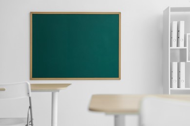 Photo of Blank green chalkboard, desks, chairs and shelving unit in classroom