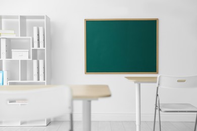 Photo of Blank green chalkboard, desks, chairs and shelving unit in classroom