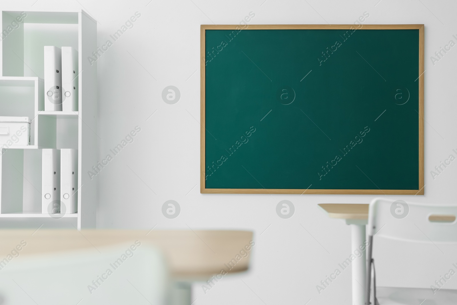 Photo of Blank green chalkboard, desks, chairs and shelving unit in classroom