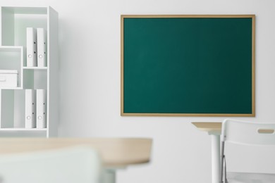 Photo of Blank green chalkboard, desks, chairs and shelving unit in classroom