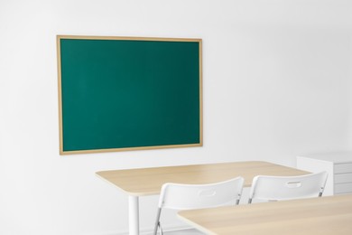 Photo of Blank green chalkboard, desks and chairs in classroom