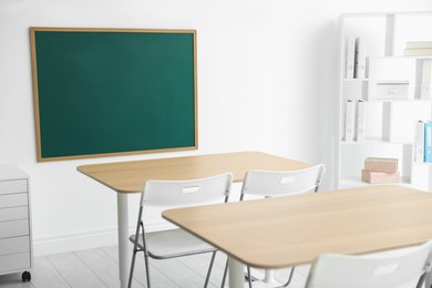 Photo of Blank green chalkboard, desks, chairs and shelving unit in classroom