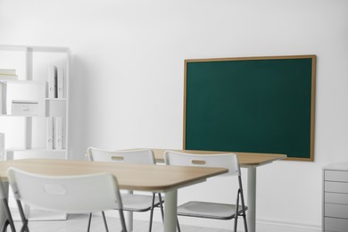 Photo of Blank green chalkboard, desks, chairs and shelving unit in classroom