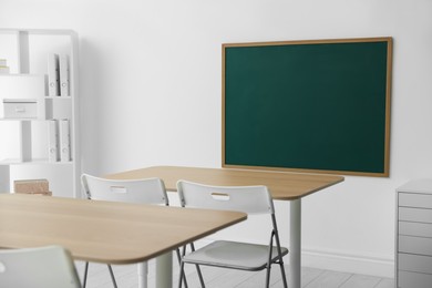 Photo of Blank green chalkboard, desks, chairs and shelving unit in classroom