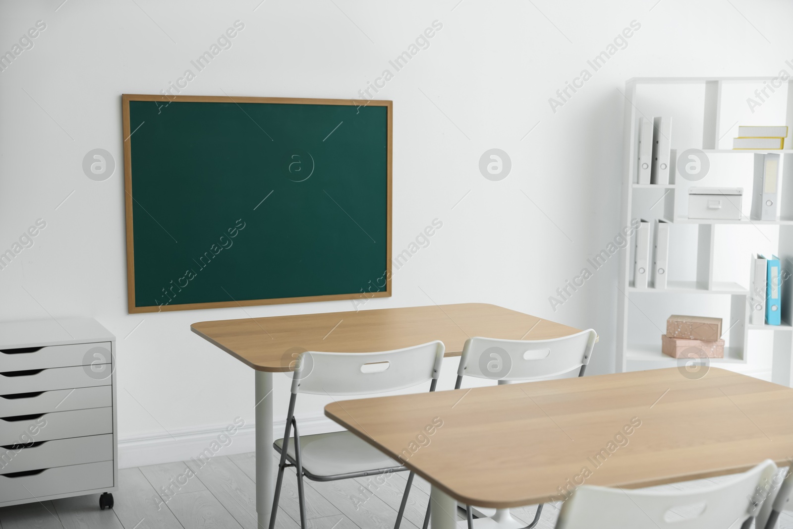 Photo of Blank green chalkboard, desks, chairs and shelving unit in classroom
