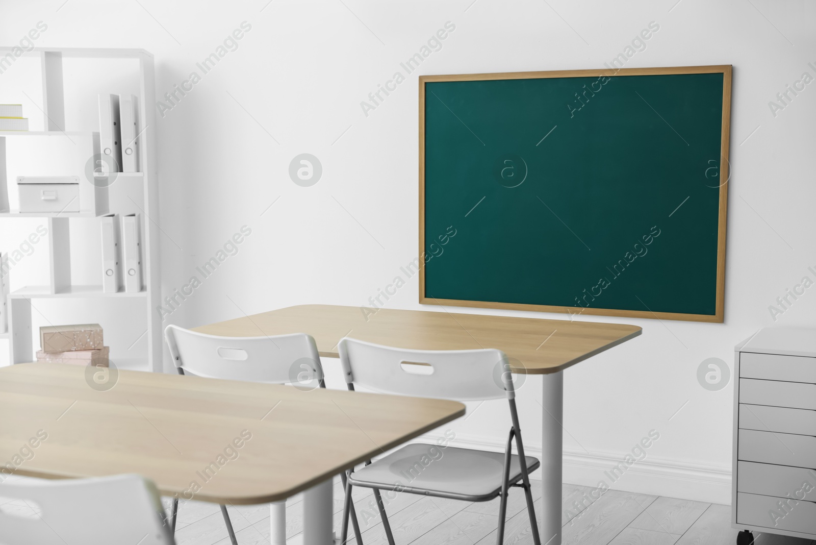 Photo of Blank green chalkboard, desks, chairs and shelving unit in classroom