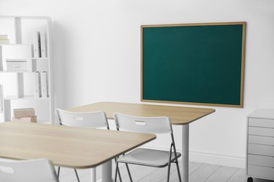 Photo of Blank green chalkboard, desks, chairs and shelving unit in classroom