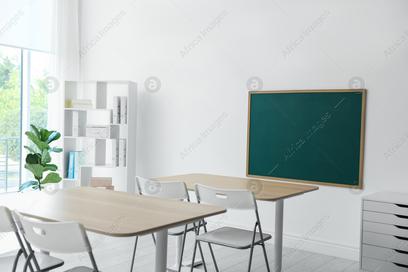 Photo of Blank green chalkboard, desks, chairs and shelving unit in classroom