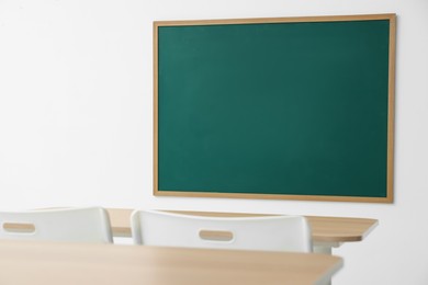 Photo of Blank green chalkboard, desks and chairs in classroom