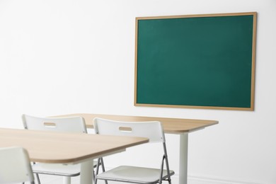 Photo of Blank green chalkboard, desks and chairs in classroom