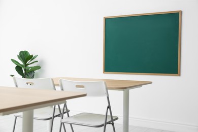 Photo of Blank green chalkboard, desks, chairs and plant in classroom