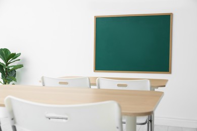 Photo of Blank green chalkboard, desks, chairs and plant in classroom