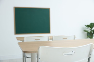 Photo of Blank green chalkboard, desks, chairs and plant in classroom, closeup
