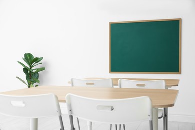 Photo of Blank green chalkboard, desks, chairs and plant in classroom