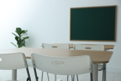 Photo of Blank green chalkboard, desks, chairs and plant in classroom, closeup