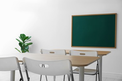 Photo of Blank green chalkboard, desks, chairs and plant in classroom