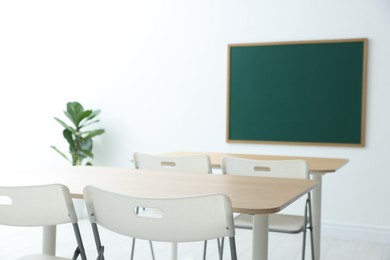 Photo of Blank green chalkboard, desks, chairs and plant in classroom, closeup