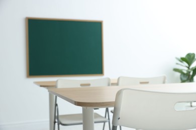 Photo of Blank green chalkboard, desks, chairs and plant in classroom, closeup