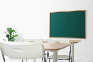 Photo of Blank green chalkboard, desks, chairs and plant in classroom