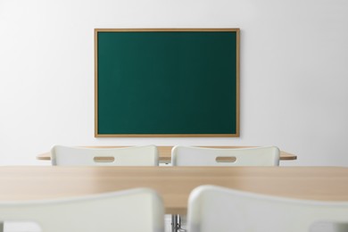 Photo of Blank green chalkboard, desks and chairs in classroom