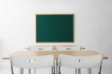 Photo of Blank green chalkboard, desks and chairs in classroom