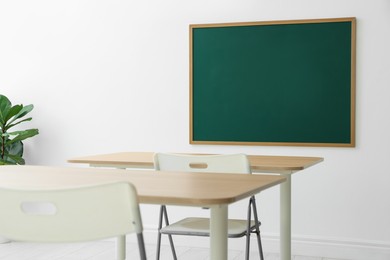 Photo of Blank green chalkboard, desks, chairs and plant in classroom