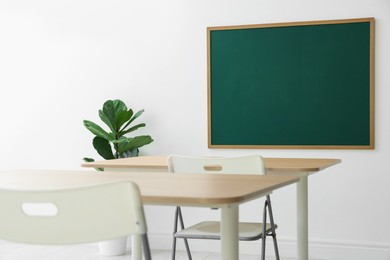 Photo of Blank green chalkboard, desks, chairs and plant in classroom