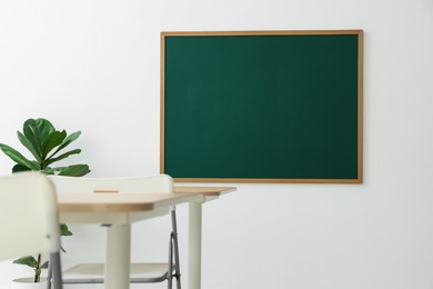 Photo of Blank green chalkboard, desks, chairs and plant in classroom