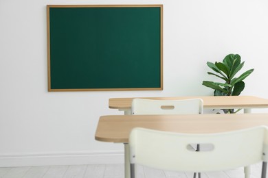 Photo of Blank green chalkboard, desks, chairs and plant in classroom