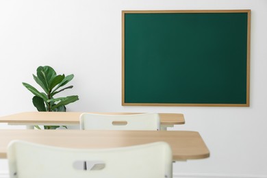 Photo of Blank green chalkboard, desks, chairs and plant in classroom