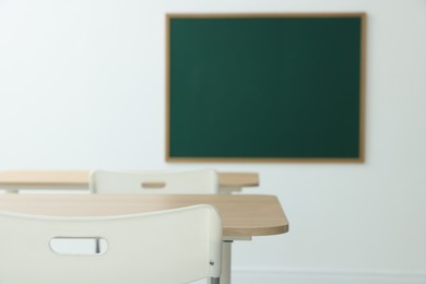 Photo of Blank green chalkboard, desks and chairs in classroom, closeup