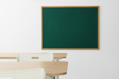 Photo of Blank green chalkboard, desks and chairs in classroom