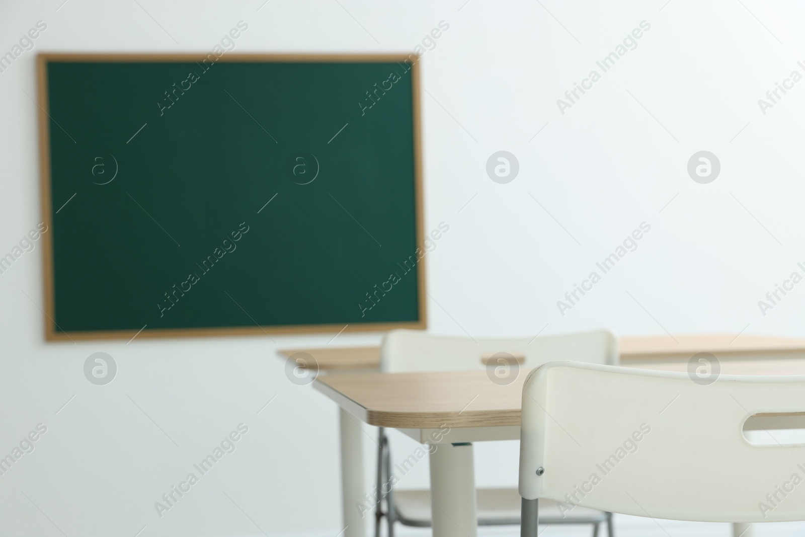 Photo of Blank green chalkboard, desks and chairs in classroom, closeup