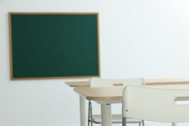 Photo of Blank green chalkboard, desks and chairs in classroom, closeup