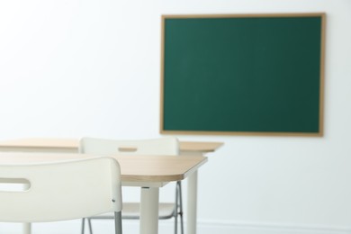 Photo of Blank green chalkboard, desks and chairs in classroom, closeup