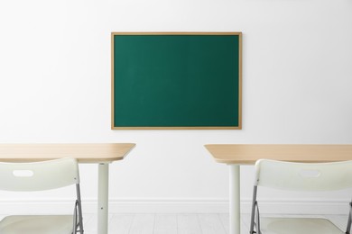 Photo of Blank green chalkboard, desks and chairs in classroom