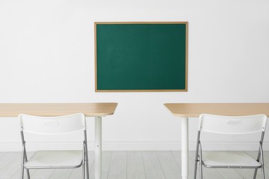 Photo of Blank green chalkboard, desks and chairs in classroom