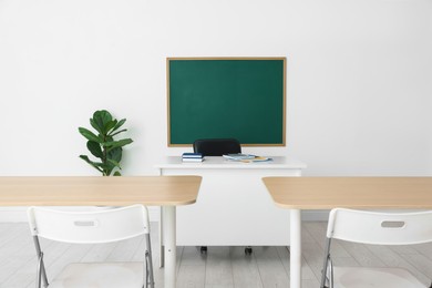 Photo of Blank green chalkboard, desks, chairs and plant in classroom