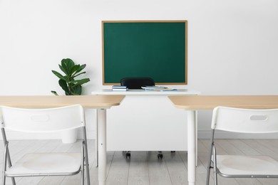 Photo of Blank green chalkboard, desks, chairs and plant in classroom