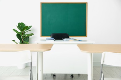 Photo of Blank green chalkboard, desks, chairs and plant in classroom