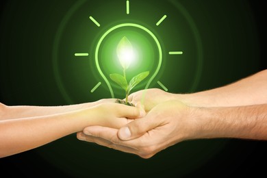 Image of Electricity and nature. Man and child holding glowing light bulb with green plant against black background, closeup