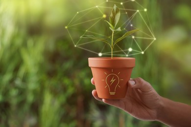 Image of Electricity and nature. Man holding potted plant against blurred green background, closeup. Light bulb with lightning symbol on pot